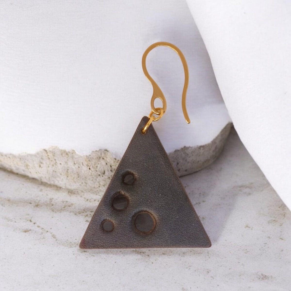 Boucle d'oreille forme triangulaire en cuir teinture noire, finition lisse et ses trous, vue de face, suspendue à une boucle dorée, fond béton