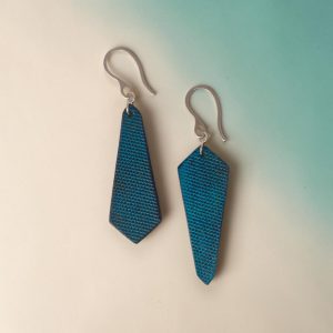 Boucles d'oreilles asymétriques en cuir teinte bleue, vues de face, suspendues à des boucles argentées fond dégradé