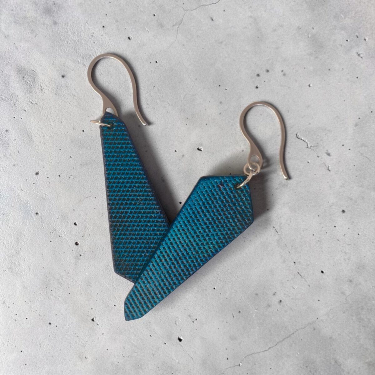 Boucles d'oreilles asymétriques en cuir teinte bleue, vues de face se chevauchant, suspendues à des boucles argentées sur béton