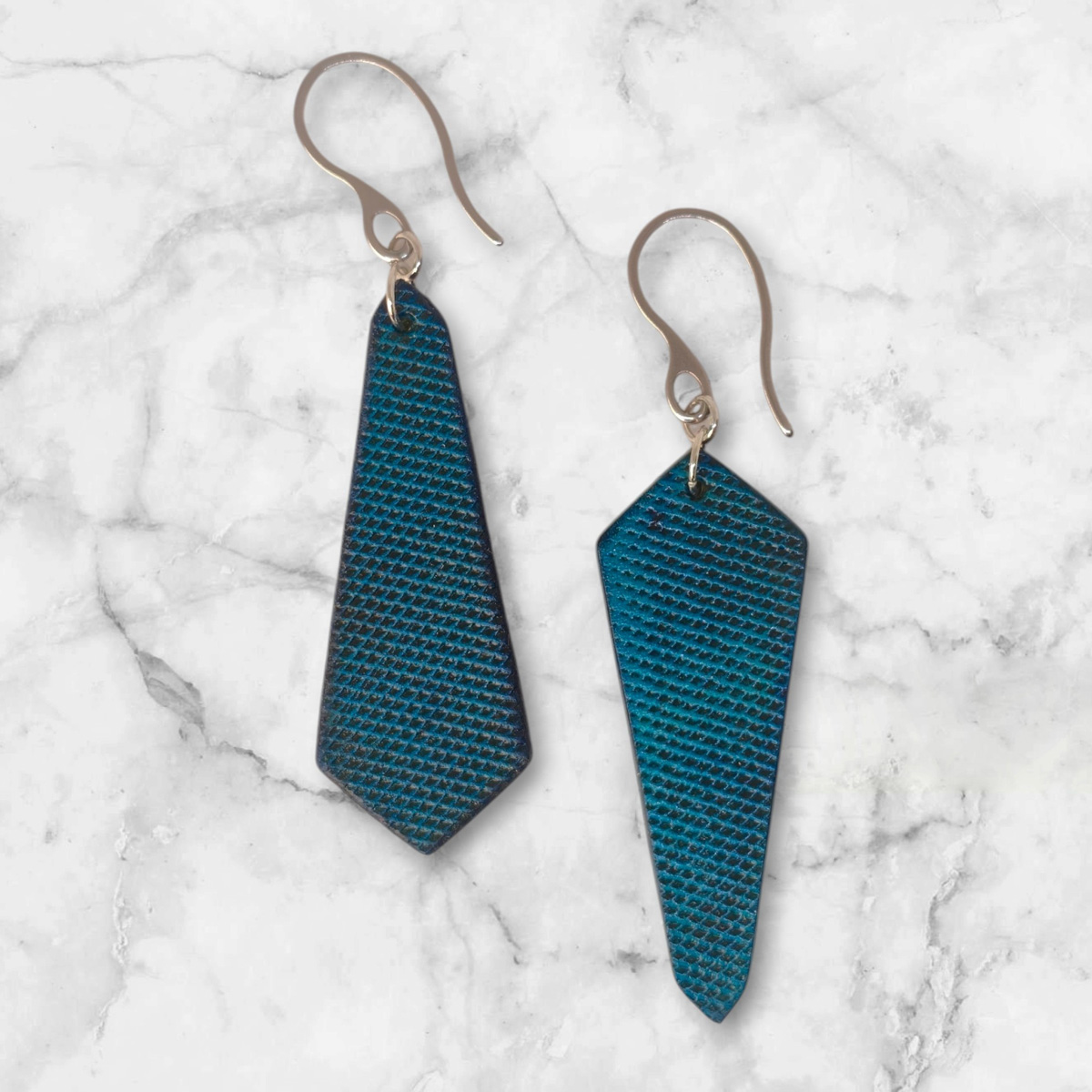 Boucles d'oreilles asymétriques en cuir teinte bleue, vues de face, suspendues à des boucles argentées posées sur marbre