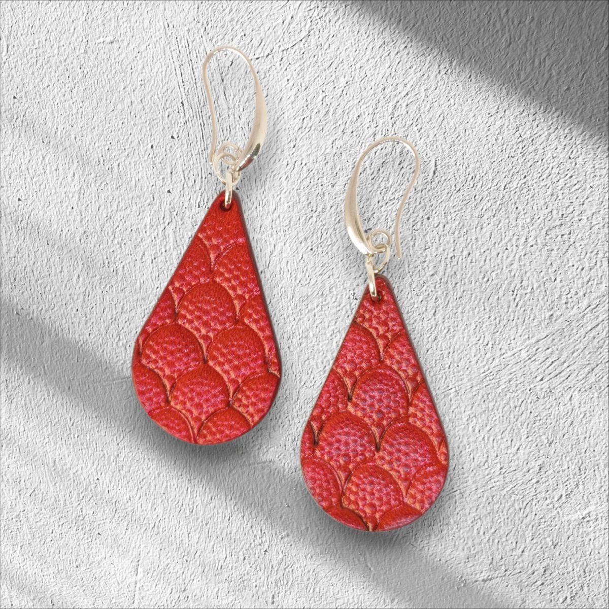 Boucles d'oreilles forme goutte d'eau en cuir teinture rouge, estampages au matoir écailles de dragon, vues de face, suspendues à des boucles argentées sur crépi
