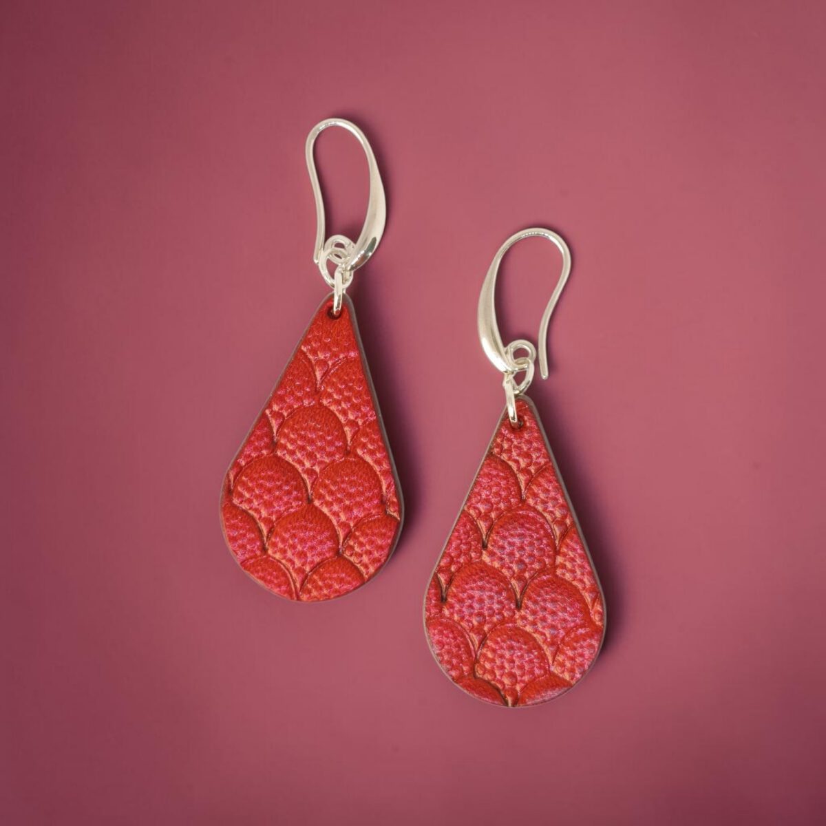 Boucles d'oreilles forme goutte d'eau en cuir teinture rouge, estampages au matoir écailles de dragon, vues de face, suspendues à des boucles argentées sur fond dégradé