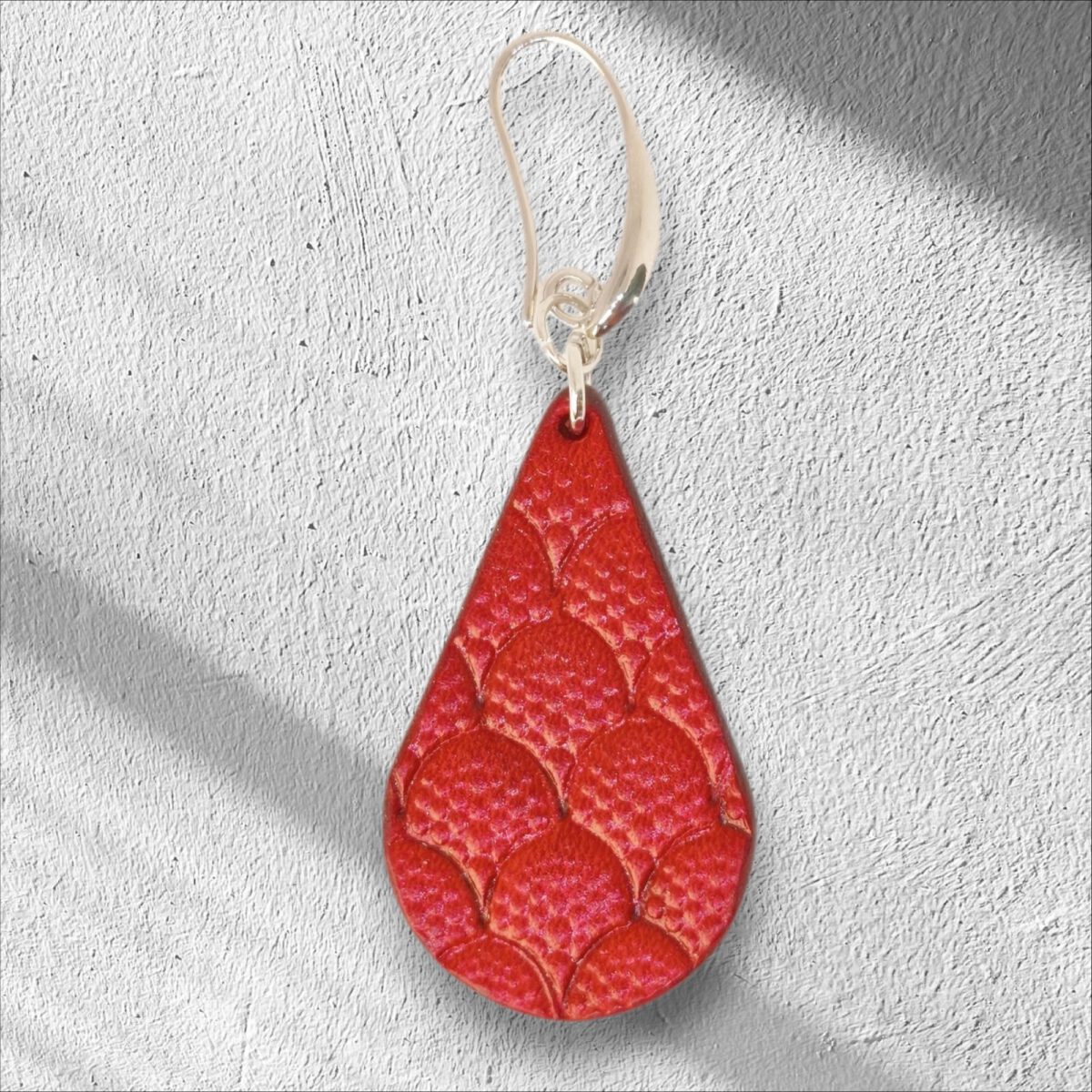 Boucle d'oreille forme goutte d'eau en cuir teinture rouge, estampage au matoir écailles de dragon, vue de face, suspendue à une boucle argentée, sur crépi