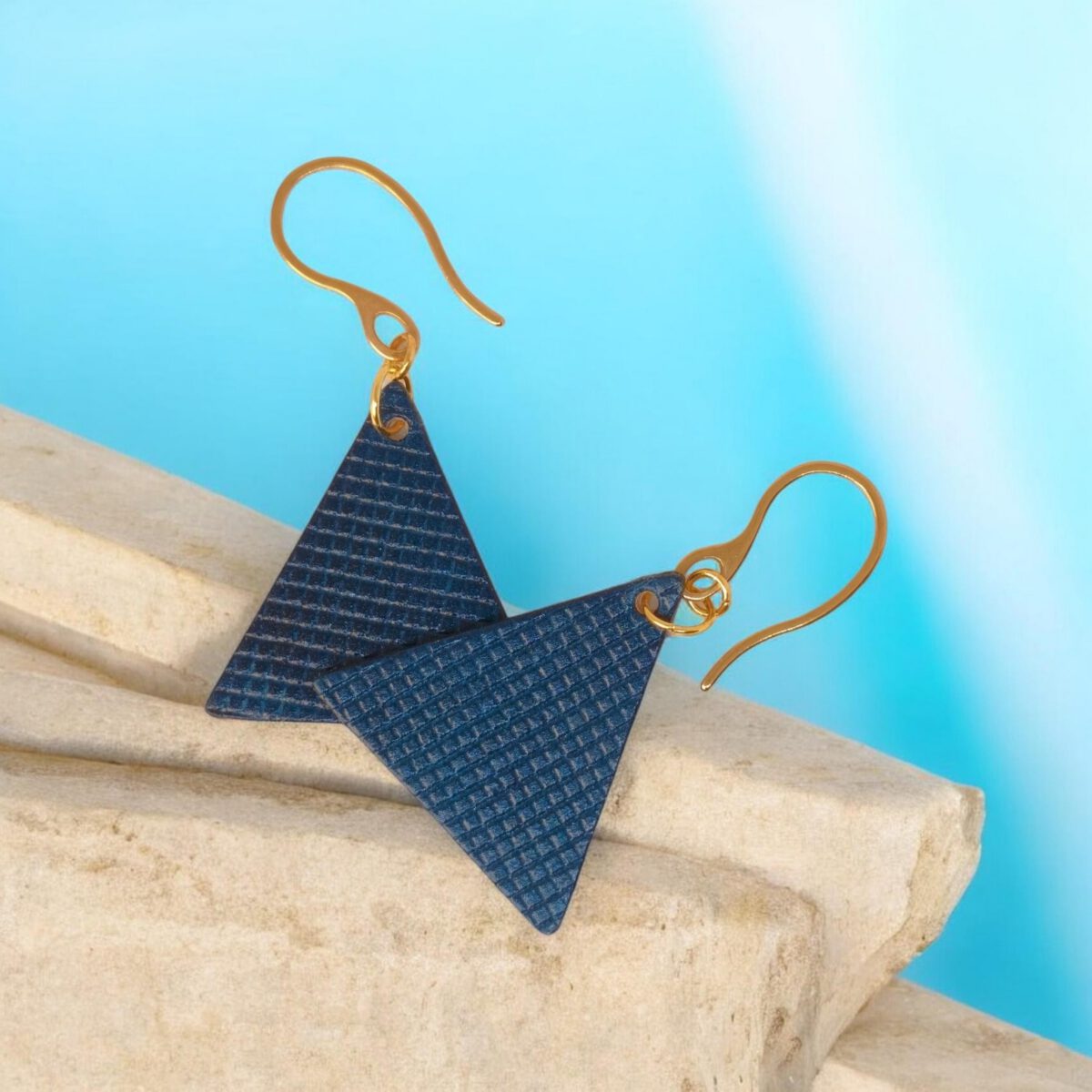Boucles d'oreilles forme triangulaire en cuir teinture bleue, texturage quadrillage, vues de face se chevauchant, suspendues à des boucles dorées sur pierre