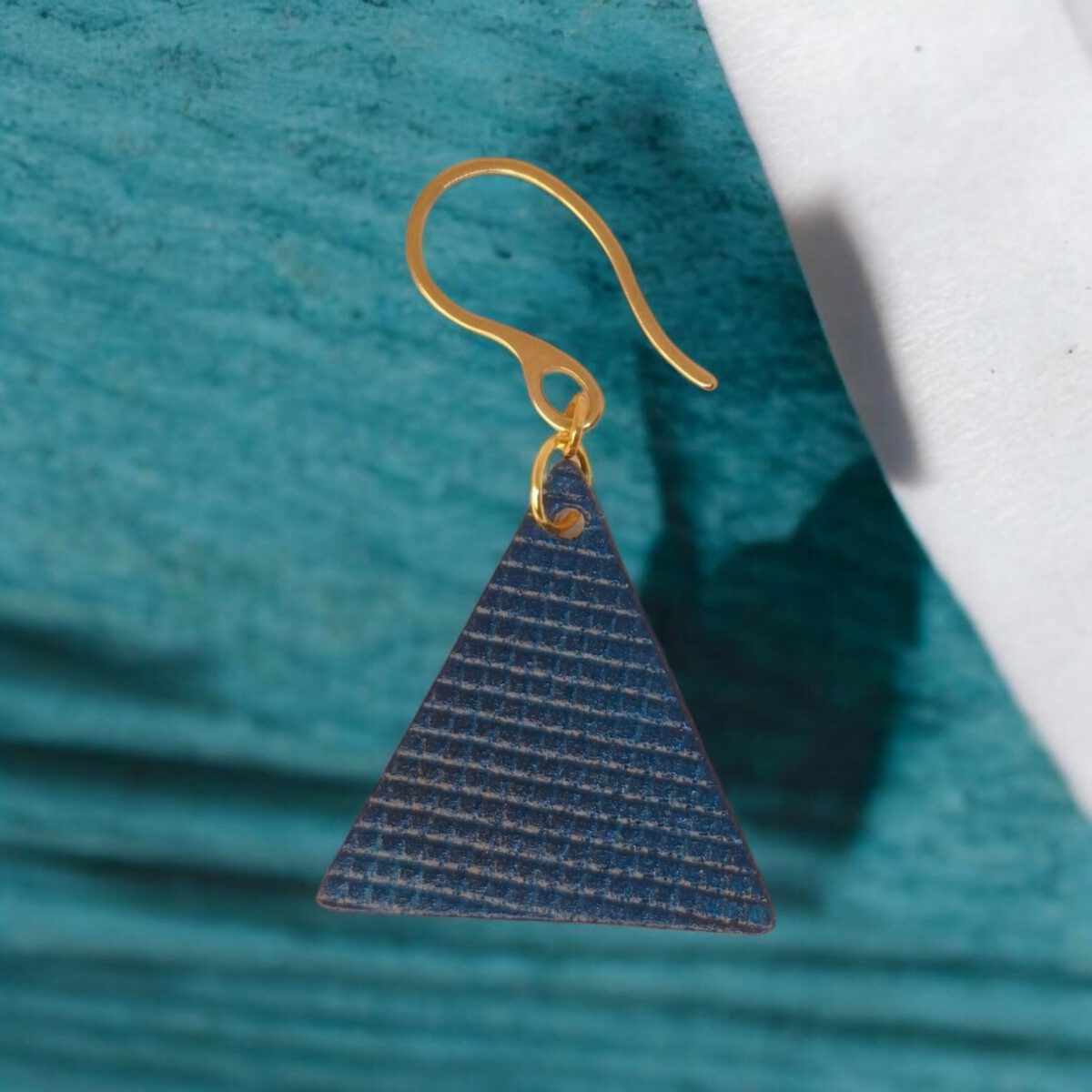 Boucles d'oreilles forme triangulaire en cuir teinture bleue, texturage quadrillage, vues de face, suspendues à des boucles dorées sur fond bleu