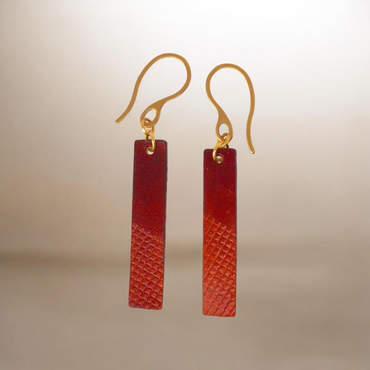 Boucles d'oreilles rectangulaires en cuir teinté cognac et acajou, finition mi-lisse mi-quadrillage , vues de face, suspendues à des boucles dorées sur fond design