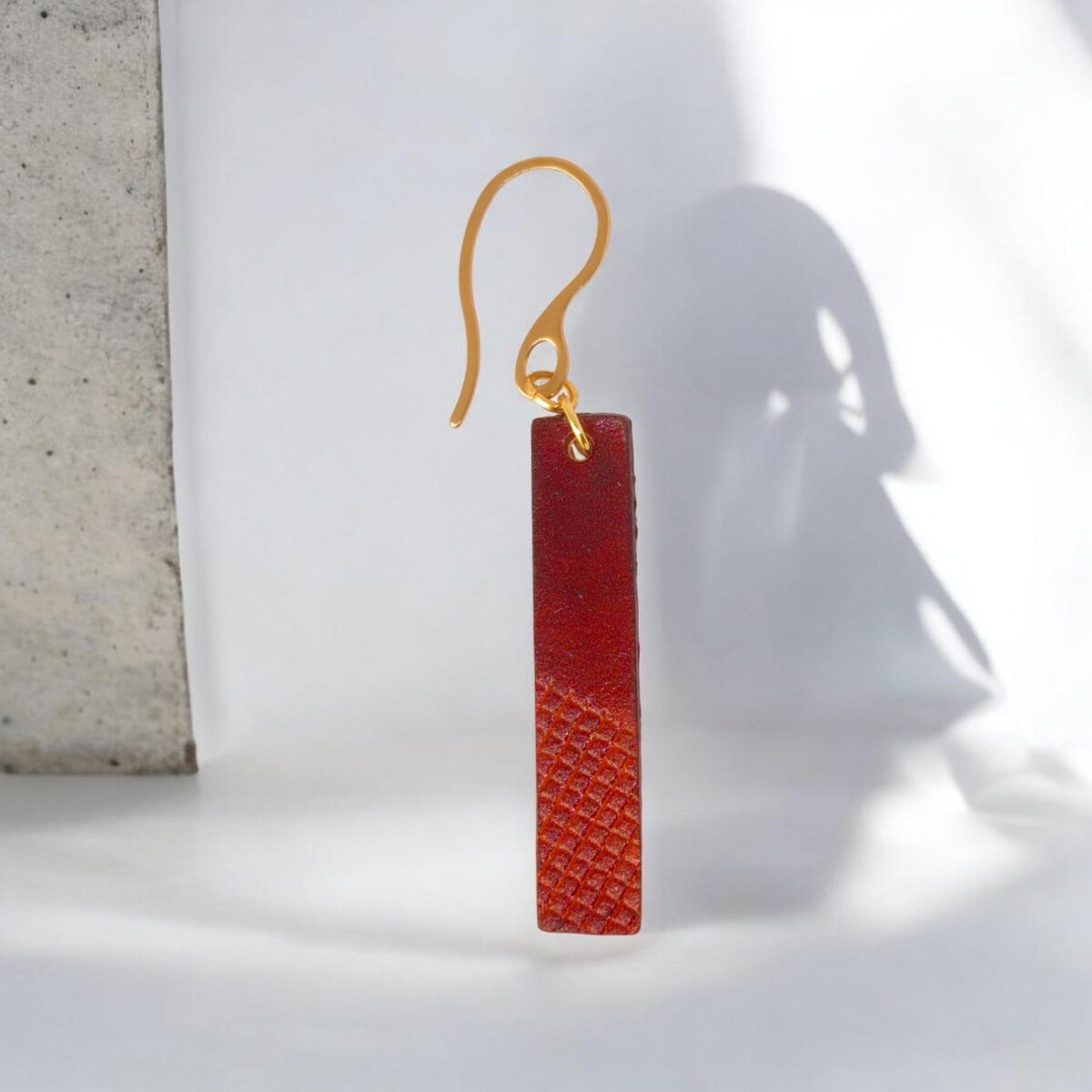Boucle d'oreille rectangulaire en cuir teinté cognac et acajou, finition mi-lisse mi-quadrillage, vue de face, suspendue à une boucle dorée, fond design