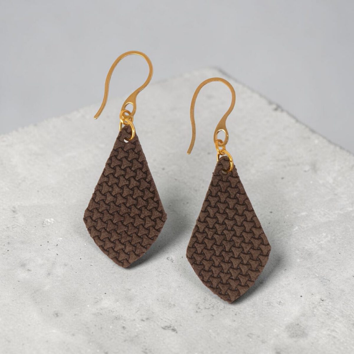 Boucles d'oreilles forme larme en cuir texturé, vues de face, suspendues à des boucles dorées, fond béton