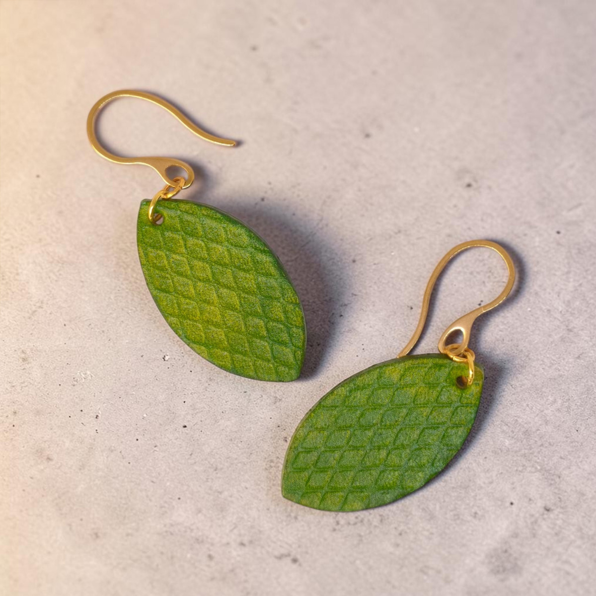Boucles d'oreilles forme poisson en cuir teinture vert forêt, texturage gros quadrillage, vues de face, suspendues à des boucles dorées sur béton