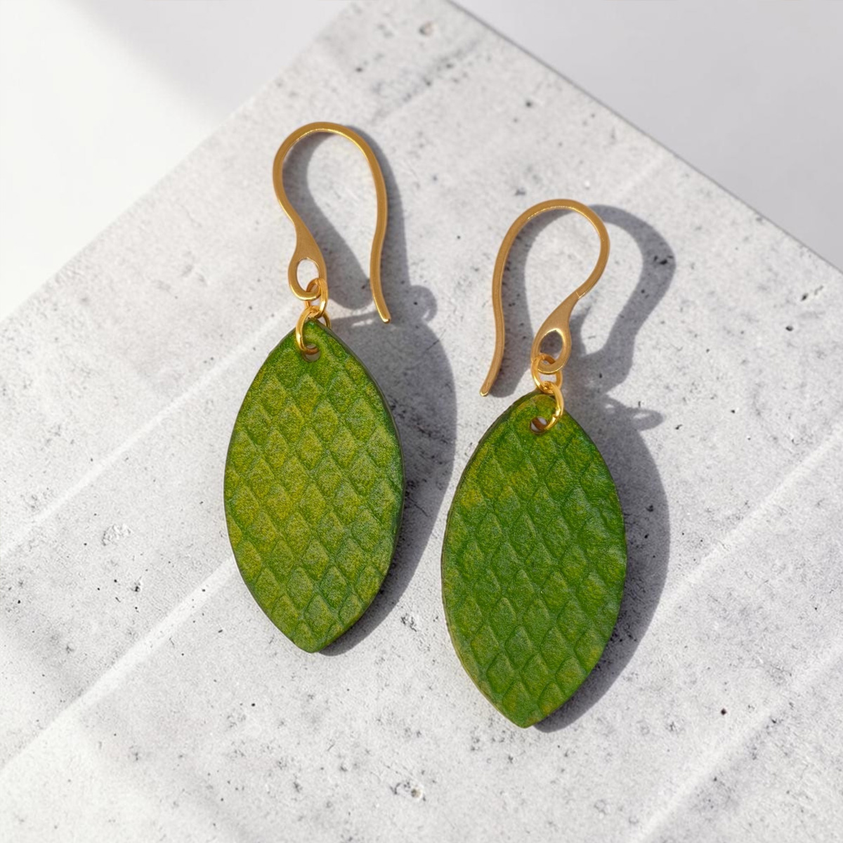 Boucles d'oreilles forme poisson en cuir teinture vert forêt, texturage gros quadrillage, vues de face, suspendues à des boucles dorées sur pierre
