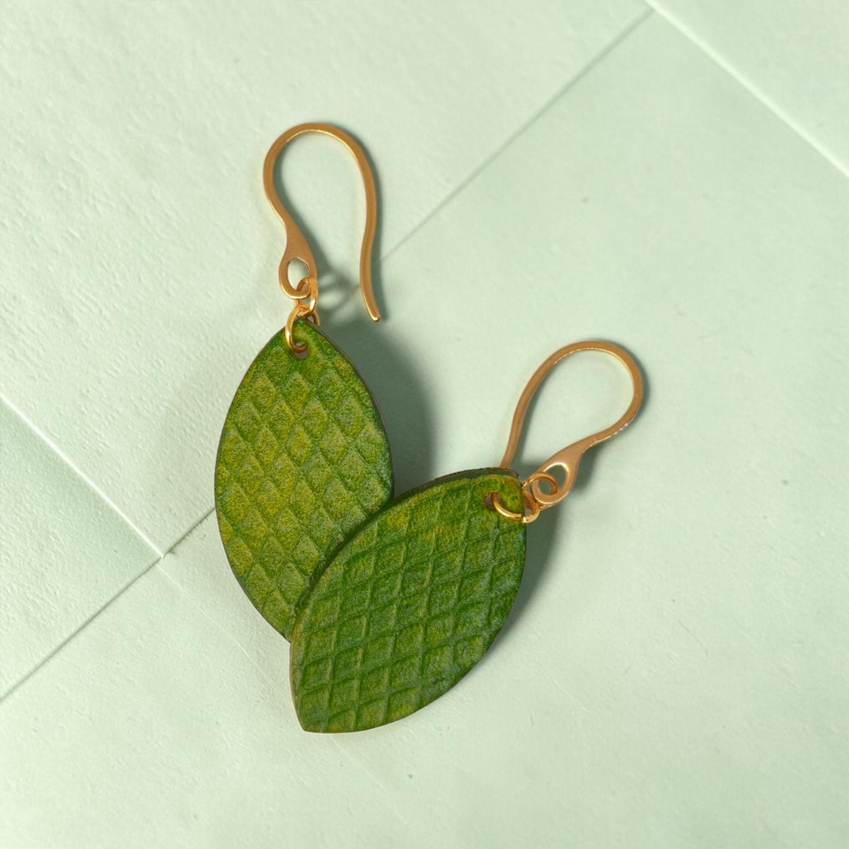 Boucles d'oreilles forme poisson en cuir teinture vert forêt, texturage gros quadrillage, vues de face, suspendues à des boucles dorées sur fond design vert