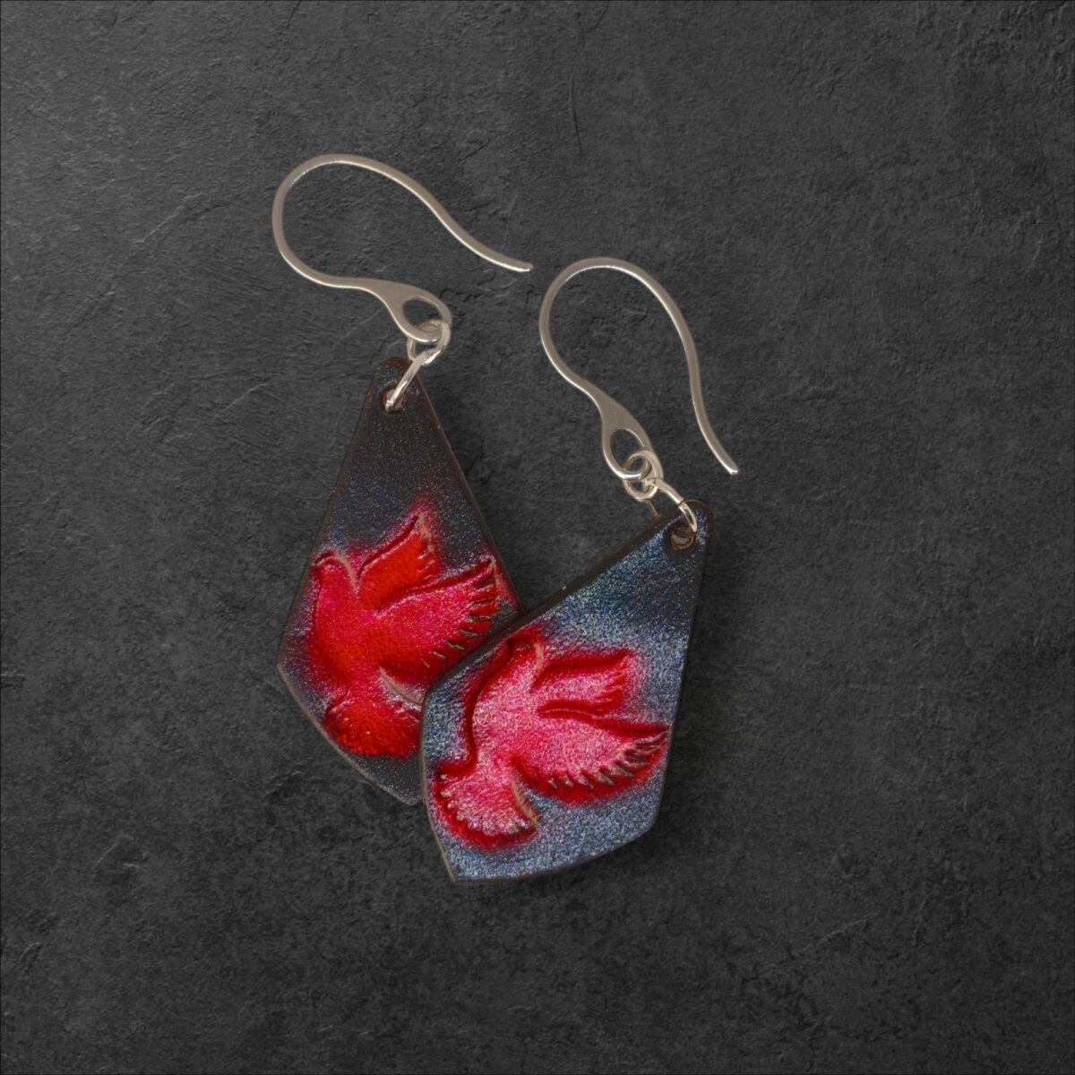 Boucles d'oreilles en cuir teintées en rouge et noir, vues de face se chevauchant, suspendues à des crochets argentés, fond noir