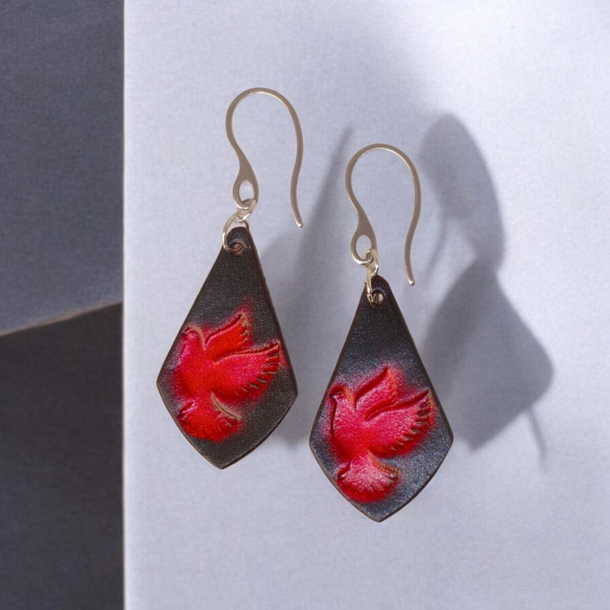 Boucles d'oreilles en cuir teintées en rouge et noir, vues de face, suspendues à des crochets argentés, colombe dégradée rouge
