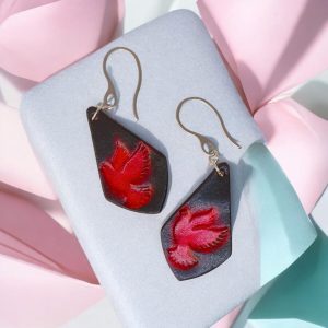 Boucles d'oreilles en cuir teintées en rouge et noir, vues de face, suspendues à des crochets argentés sur fond design