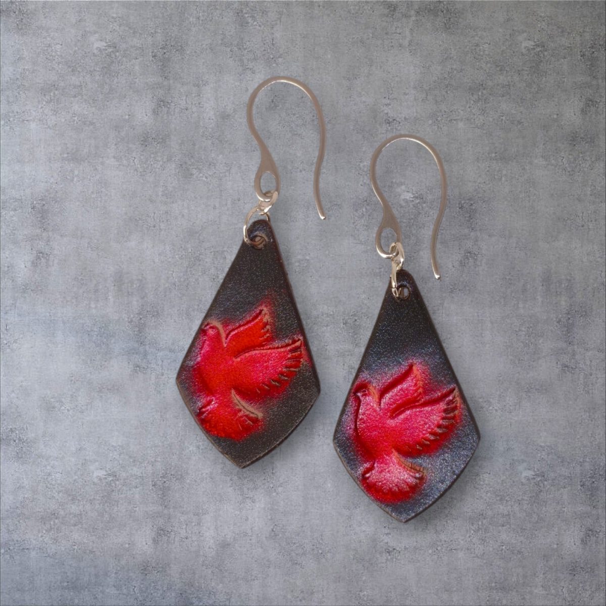 Boucles d'oreilles en cuir teintées en rouge et noir, vues de face, suspendues à des crochets argentés sur fond gris
