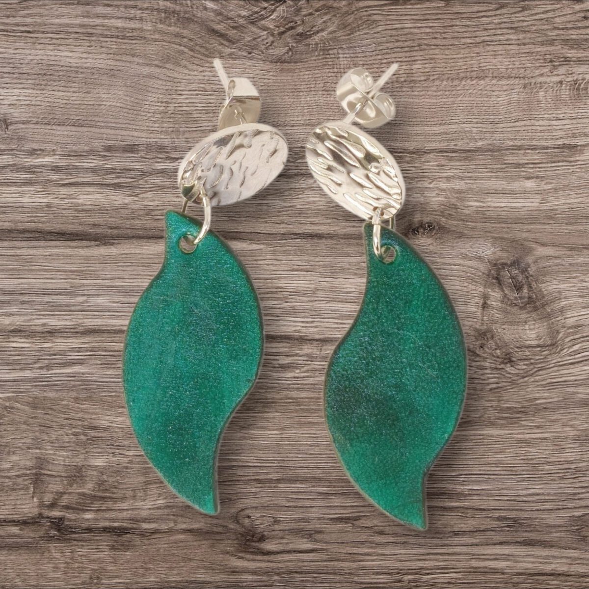 Boucles d'oreilles forme feuille asymétrique en cuir teinté vert d'eau, finition lisse, vues de face, suspendues à des boucles argentées, fond bois