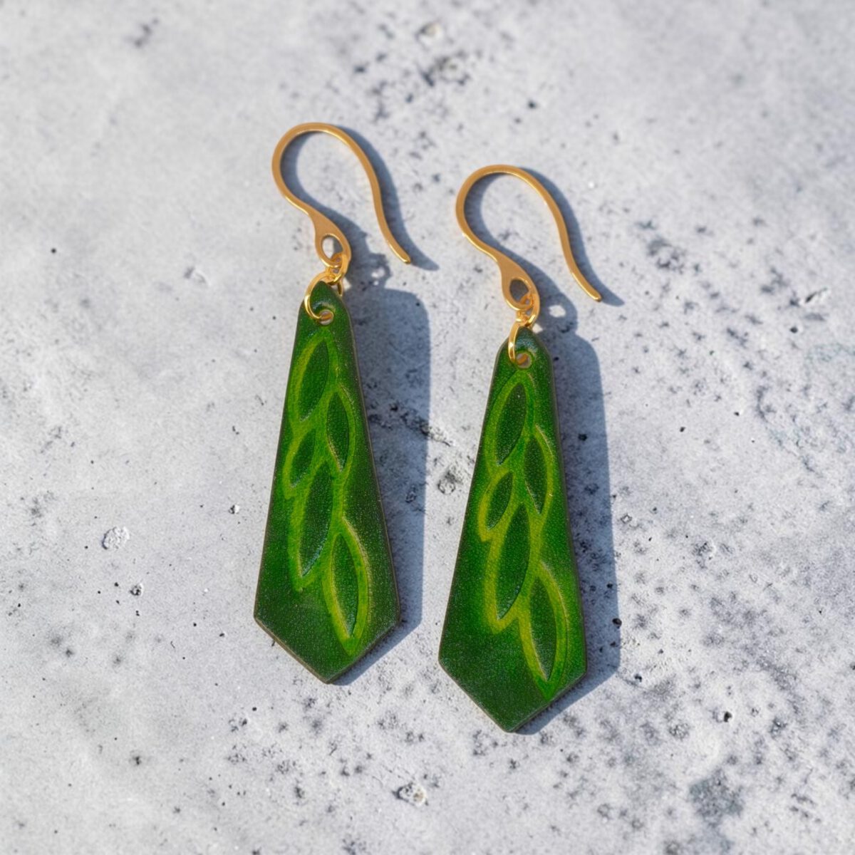 Boucles d'oreilles cravates en cuir teinté vert forêt, estampage feuille longue, vues de face, suspendues à des boucles dorées, sur béton
