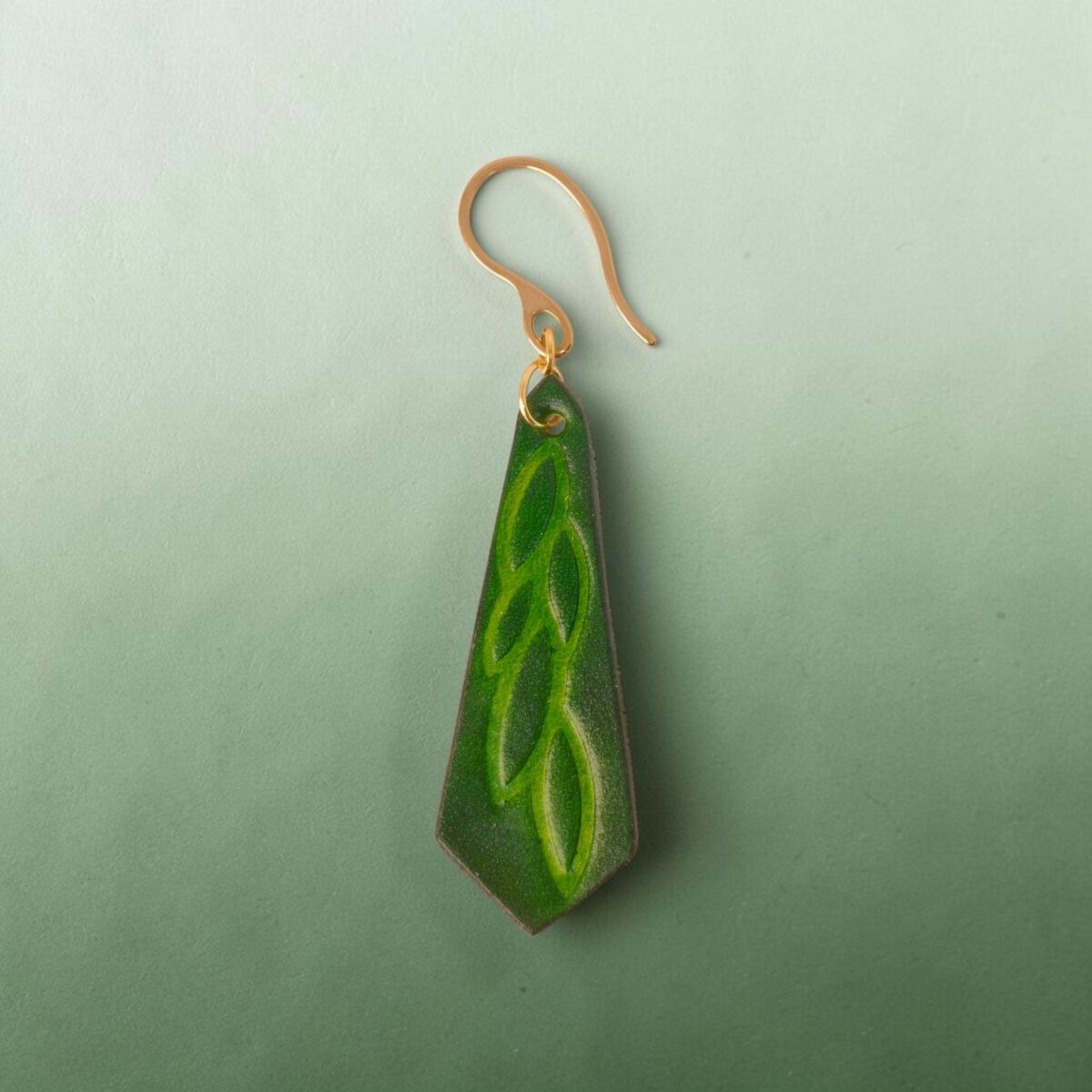 Boucle d'oreille cravate en cuir teinté vert forêt, estampage feuille longue, vue de face, suspendue à une boucle dorée, fond design