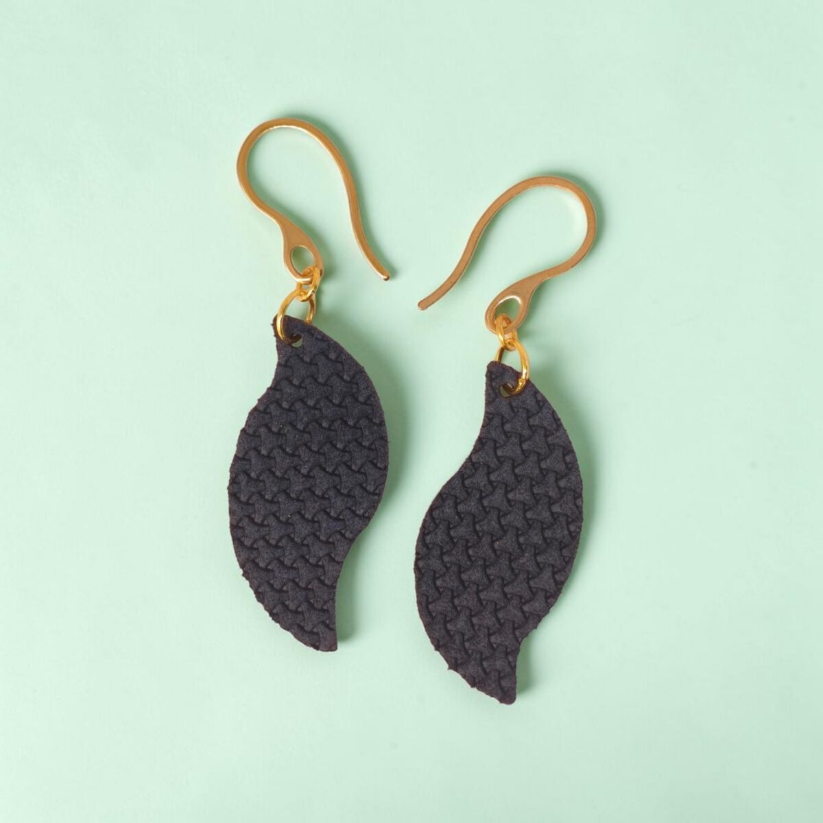Boucles d'oreilles forme feuille asymétrique en cuir teinté bleu nuit, texturage en tannerie, vues de face, suspendues à des boucles dorées sur fond design