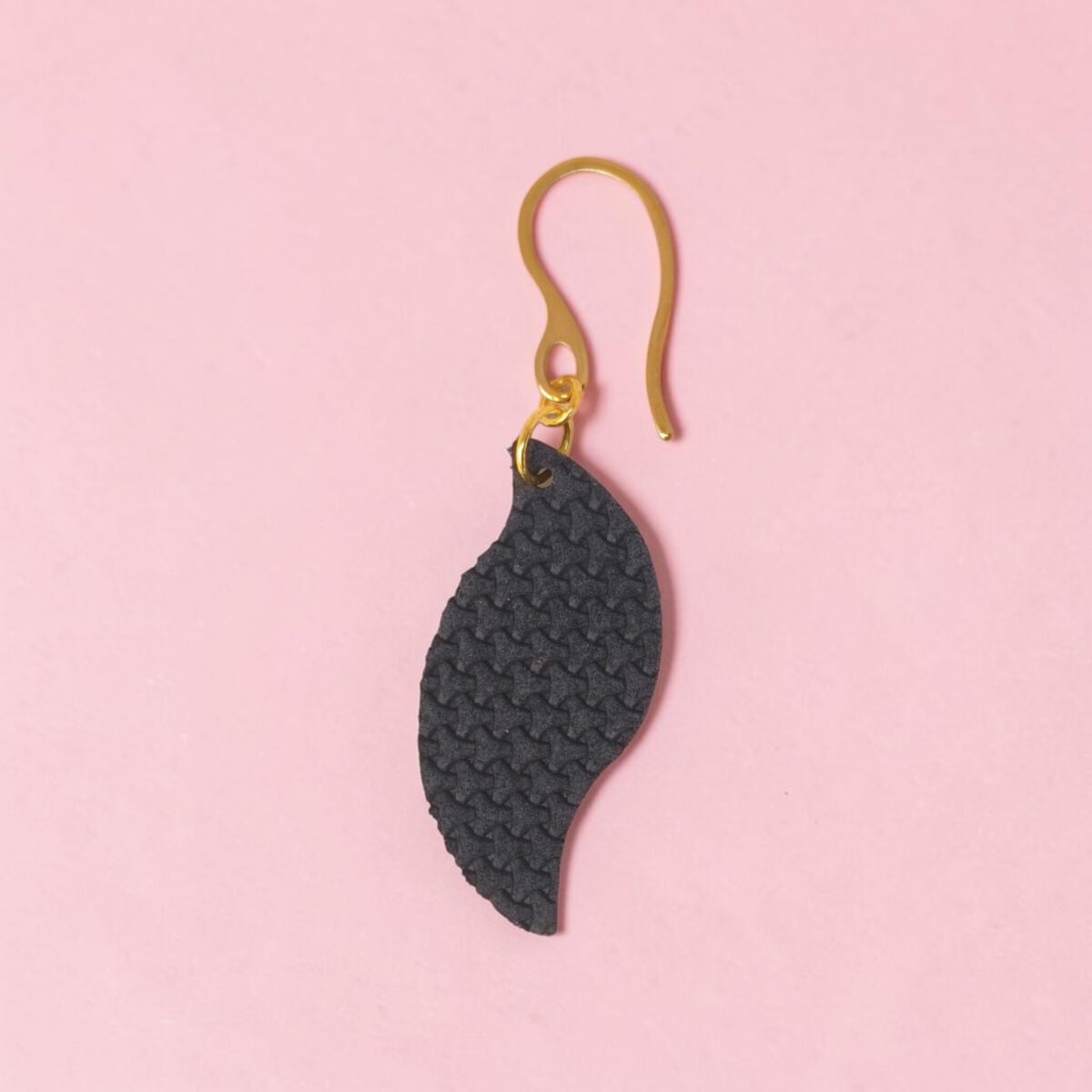 Boucle d'oreille forme feuille asymétrique en cuir teinté bleu nuit, texturage en tannerie, vue de face, suspendue à une boucle dorée, fond rose