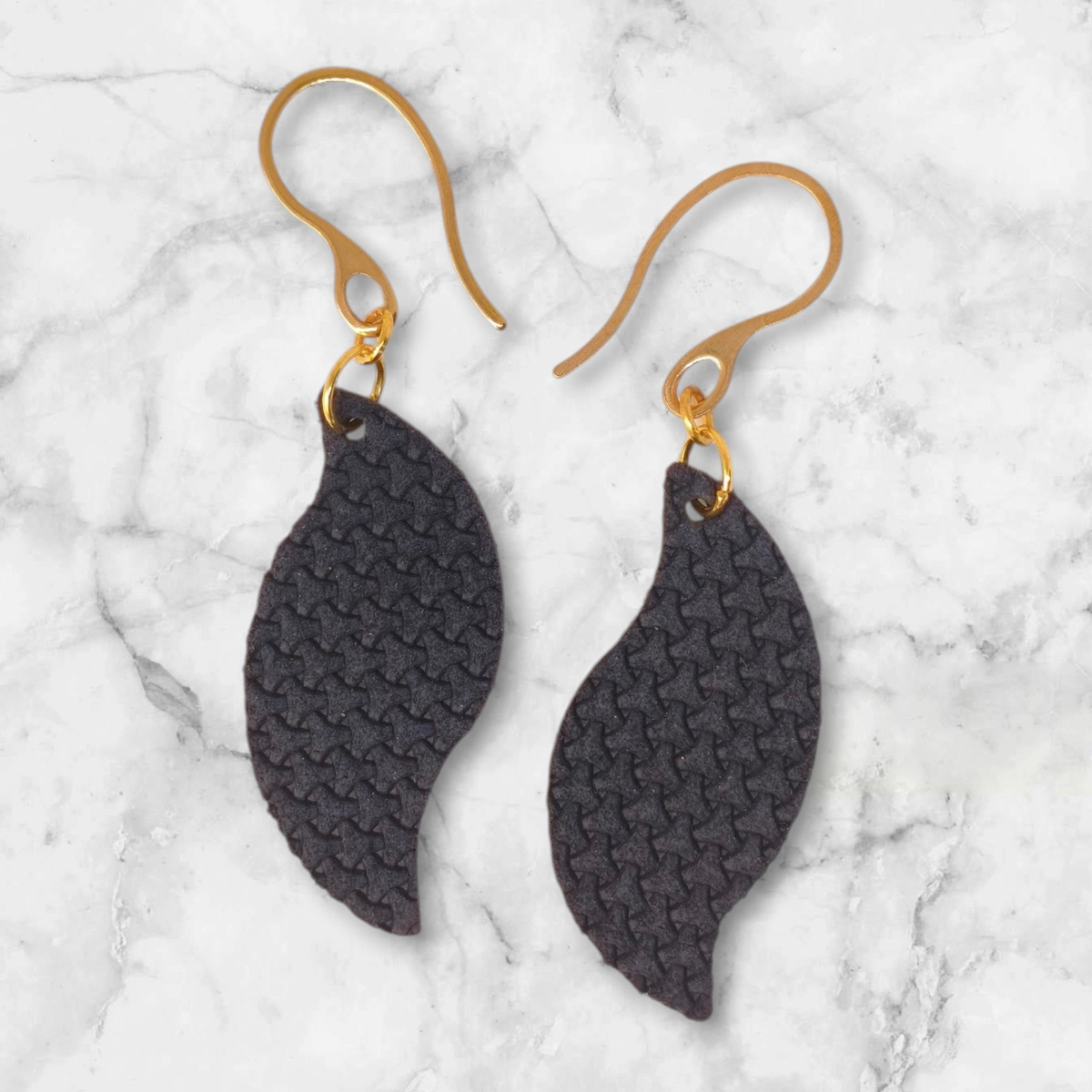 Boucles d'oreilles forme feuille asymétrique en cuir teinté bleu nuit, texturage en tannerie, vues de face, suspendues à des boucles dorées sur marbre