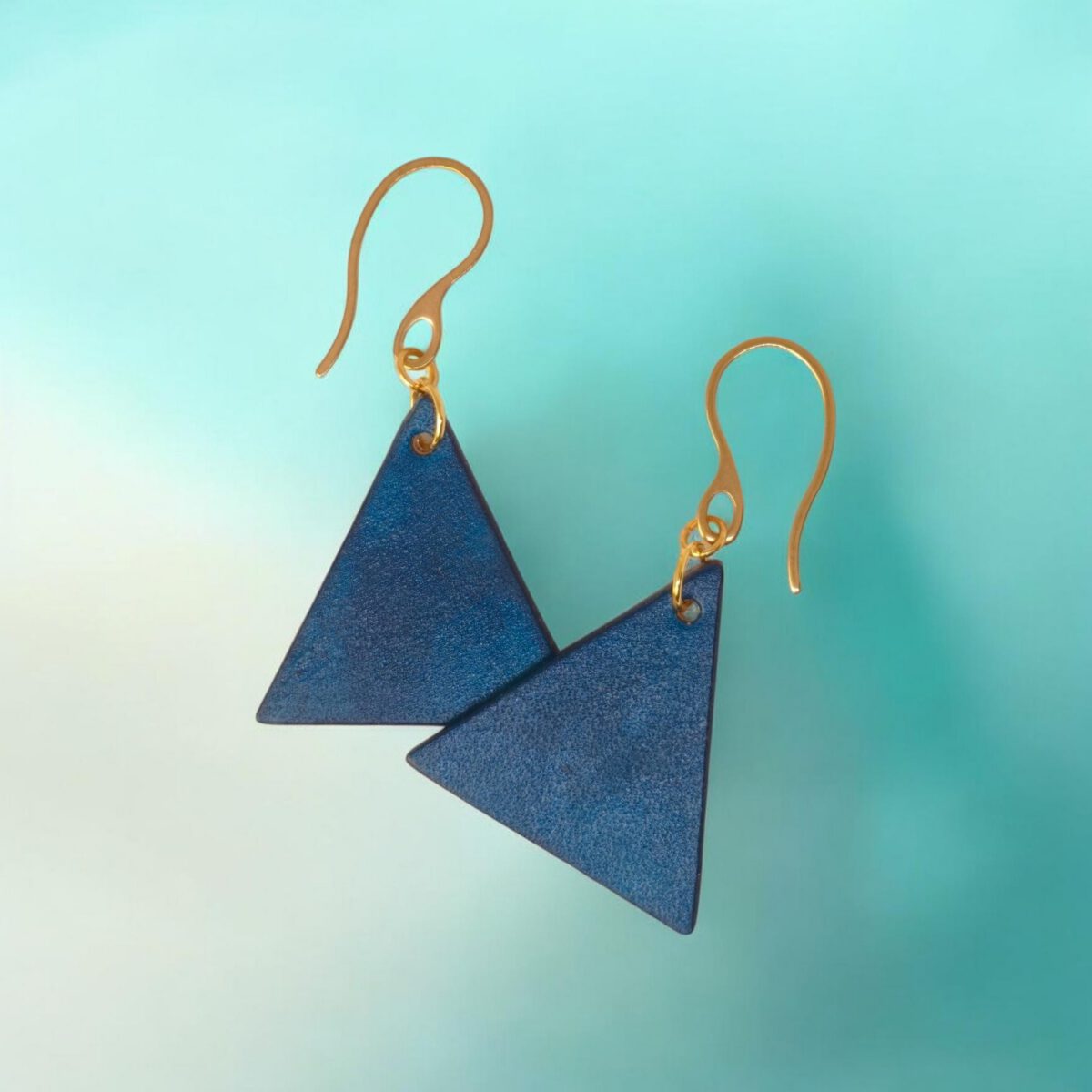 Boucles d'oreilles forme triangulaire en cuir teinture bleue, finition lisse, vues de face se chevauchant, suspendues à des boucles dorées sur fond dégradé
