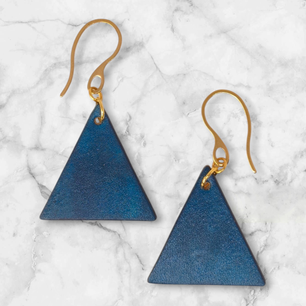 Boucles d'oreilles forme triangulaire en cuir teinture bleue, finition lisse, vues de face, suspendues à des boucles dorées sur marbre