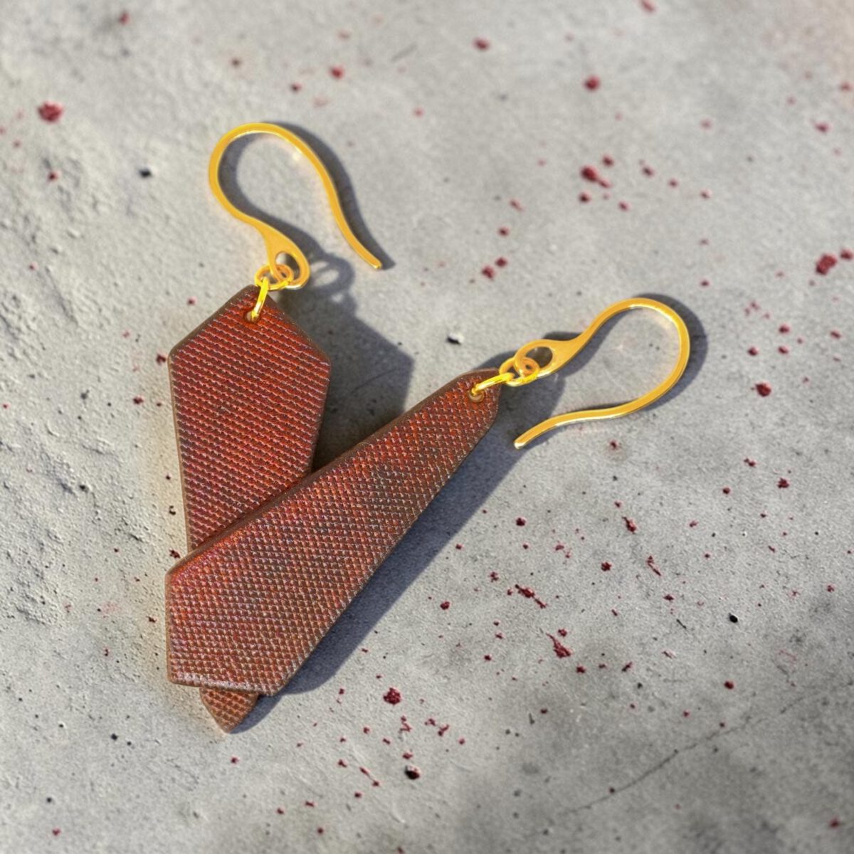 Boucles d'oreilles cravates asymétriques en cuir teinté cognac, finition lime vieillie, vues de face se chevauchant, suspendues à des boucles dorées sur fond gris