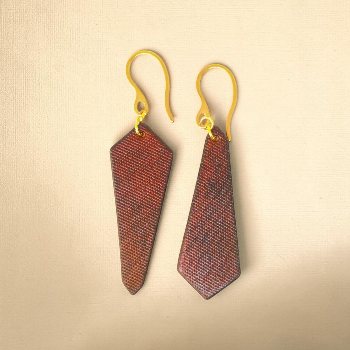 Boucles d'oreilles cravates asymétriques en cuir teinté cognac, finition lime vieillie, vues de face, suspendues à des boucles dorées sur fond degradé