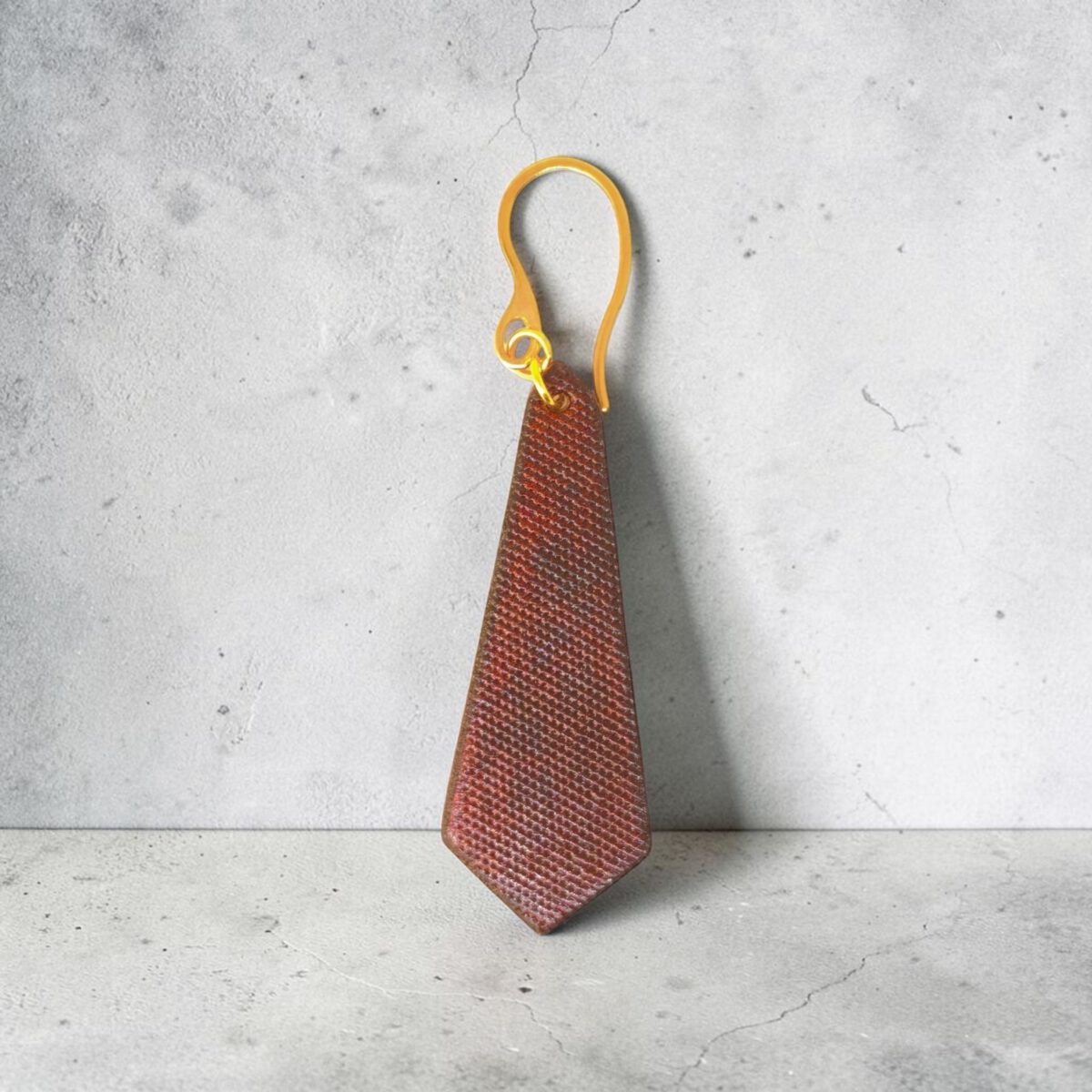Boucle d'oreille cravate asymétrique en cuir teinté cognac, finition lime vieillie, vue de face, suspendue à une boucle dorée, sur béton