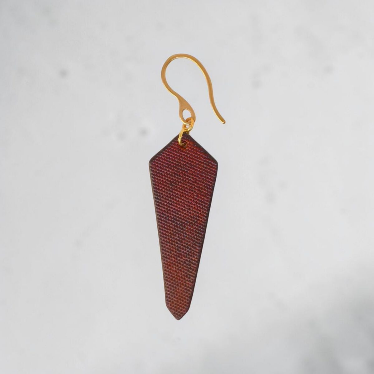 Boucle d'oreille cravate asymétrique en cuir teinté cognac, finition lime vieillie, vue de face, suspendue à une boucle dorée, fond gris flouté