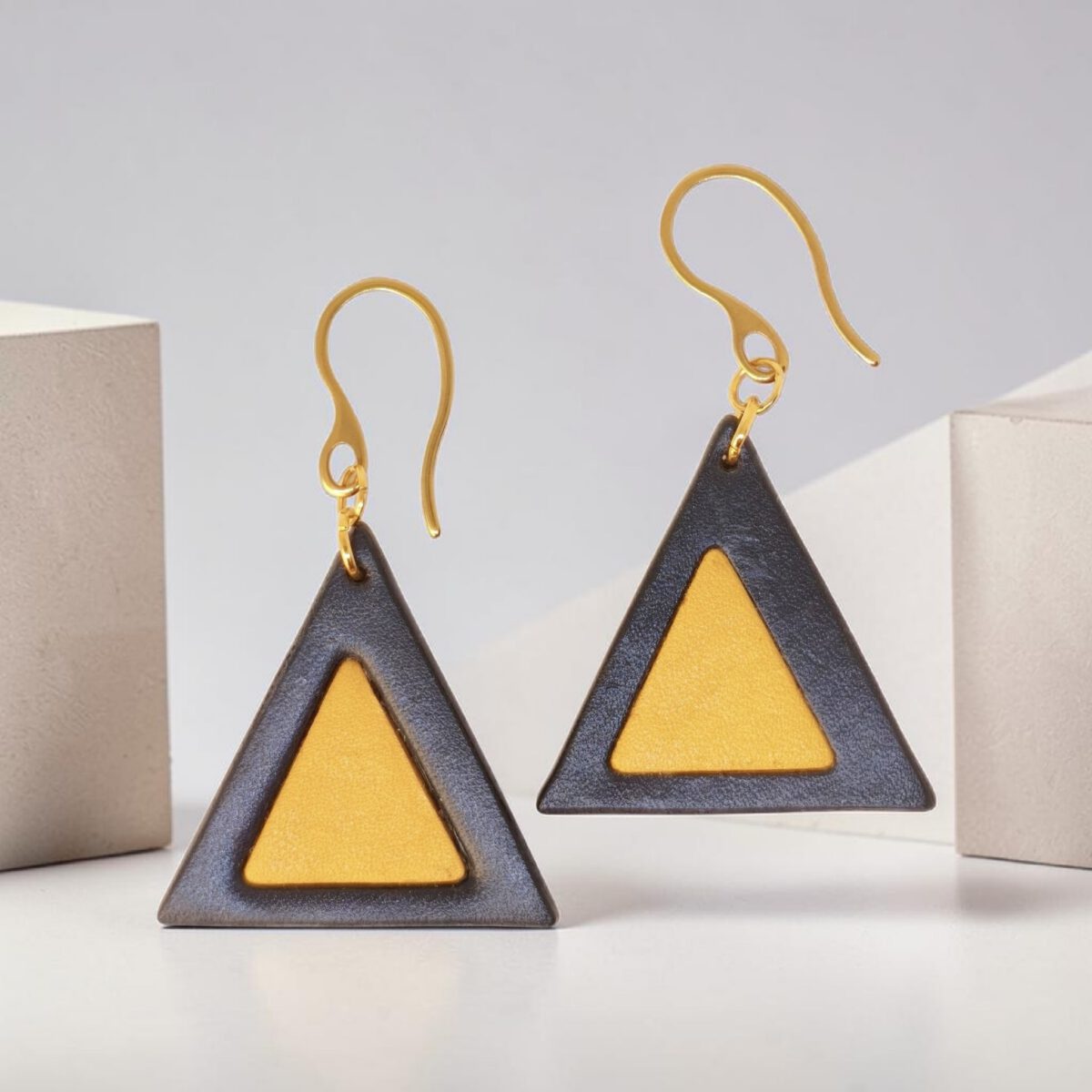 Boucles d'oreilles forme triangulaire double asymétrique en cuir teinture noire et jaune, finition lisse, vues de face, suspendues à des boucles dorées, fond design