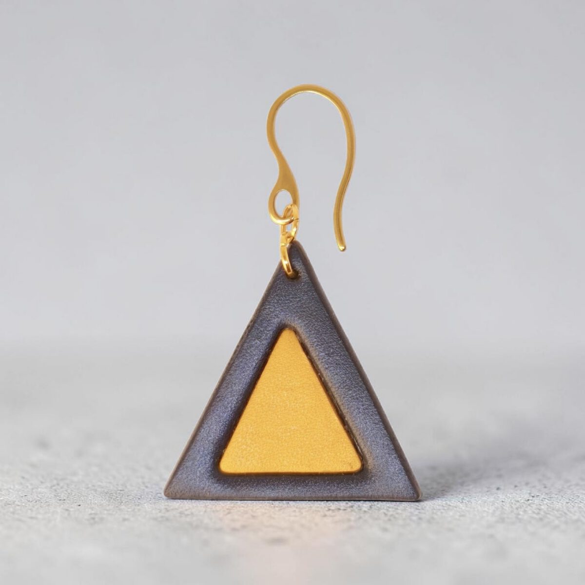 Boucle d'oreille forme triangulaire double asymétrique en cuir teinture noire et jaune, finition lisse, vue de face, suspendue à une boucle dorée, sur béton
