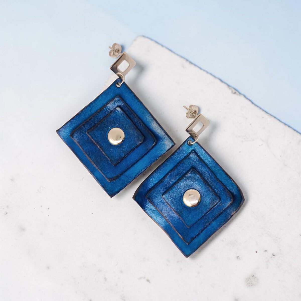 Boucles d'oreilles carrées en cuir teinté bleu, triple épaisseur, vues de face, suspendues à des boucles argentées sur pierre