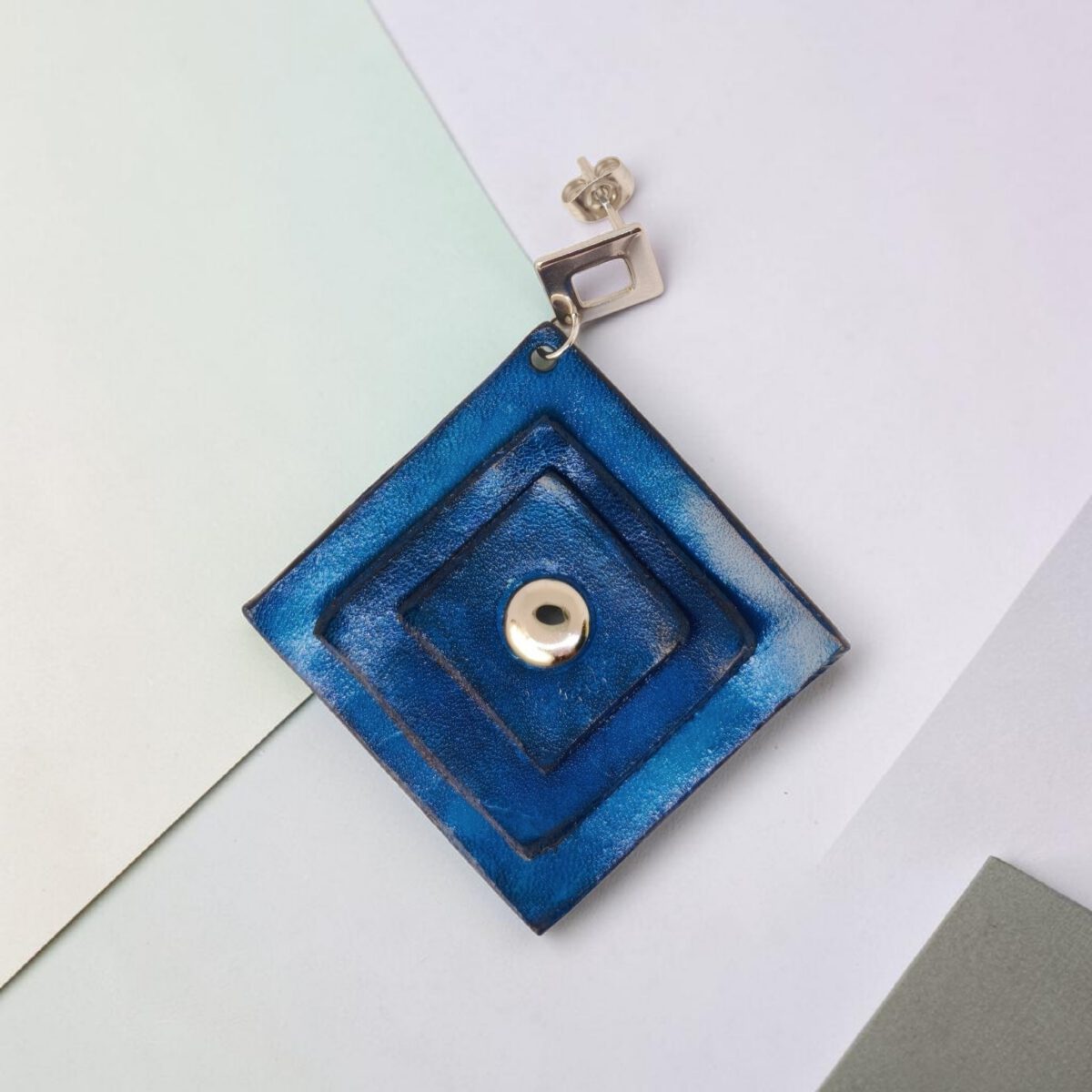 Boucle d'oreille carrée en cuir teinté bleu, triple épaisseur, vue de face, suspendue à une boucle argentée, fond design origami