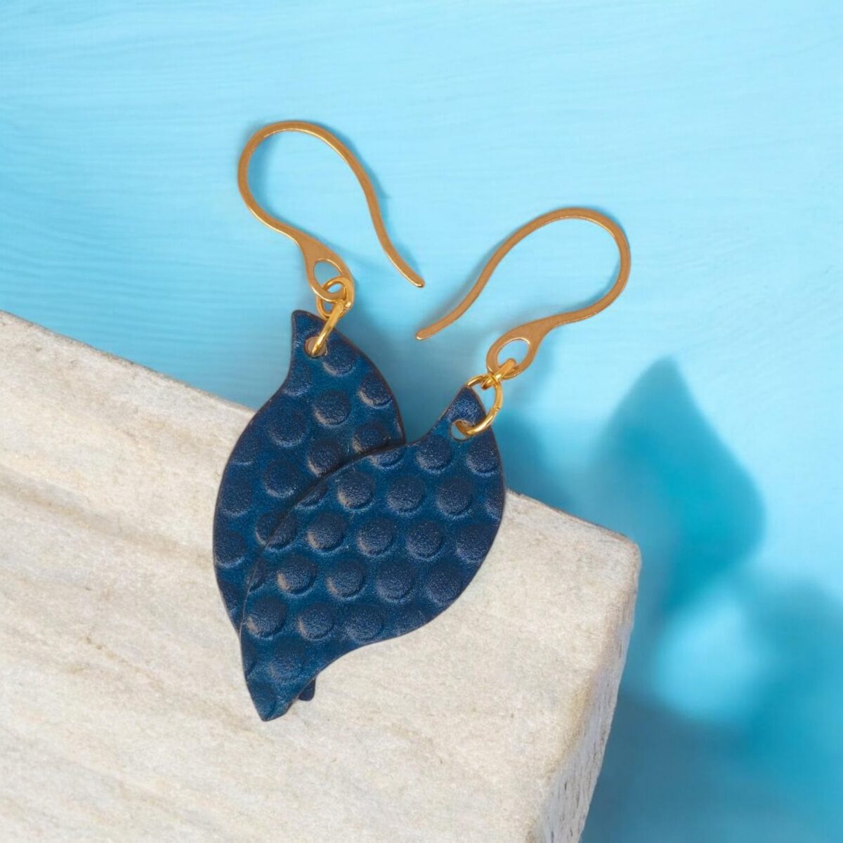Boucles d'oreilles forme feuille asymétrique en cuir teinté bleu, texturage ronds, vues de face se chevauchant, suspendues à des boucles dorées sur pierre