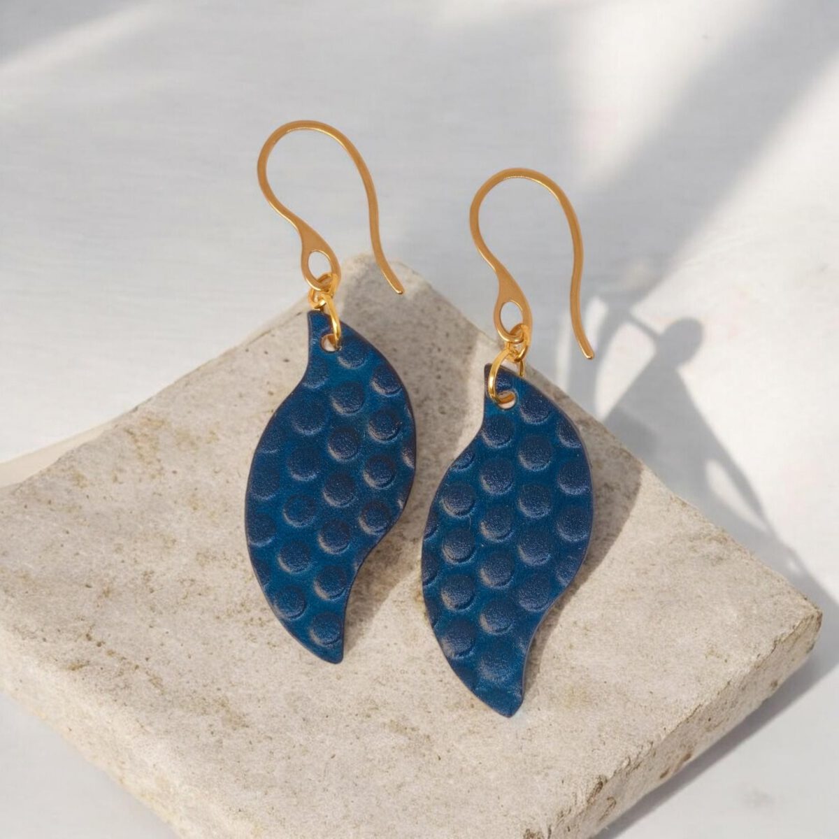 Boucles d'oreilles forme feuille asymétrique en cuir teinté bleu, texturage ronds, vues de face, suspendues à des boucles dorées sur pierre
