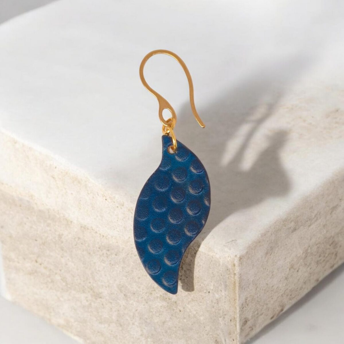 Boucle d'oreille forme feuille asymétrique en cuir teinté bleu, texturage ronds, vue de face, suspendue à une boucle dorée, sur pierre