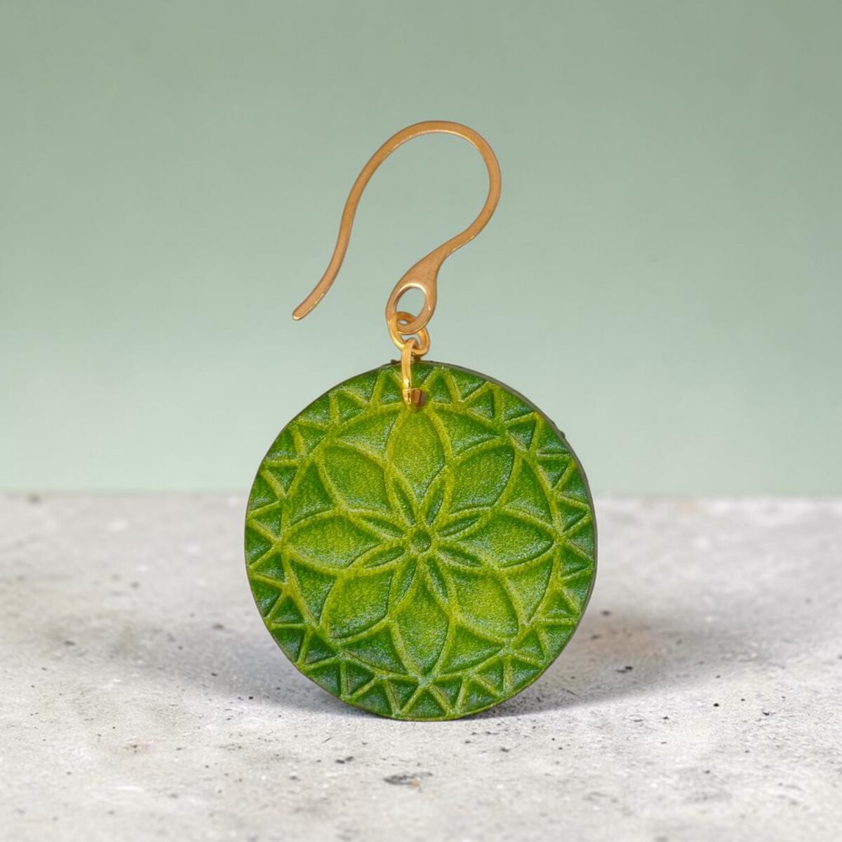 Boucle d'oreille rondes en cuir teinté vert forêt, texturage fleur mandala, vue de face, suspendue à une boucle dorée, sur béton