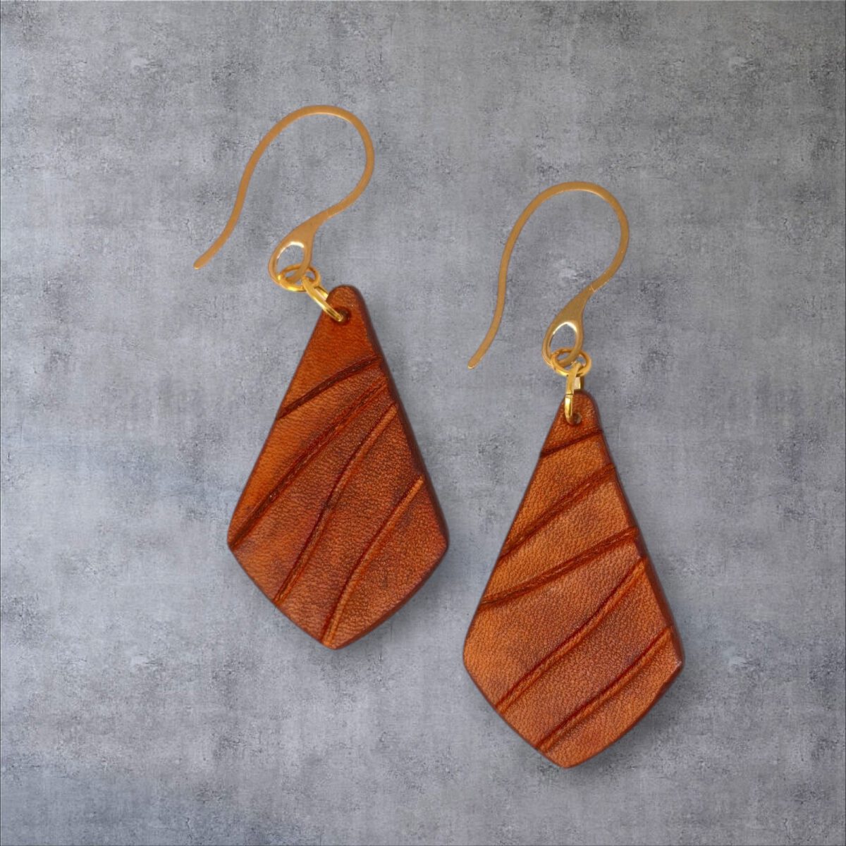 Boucles d'oreilles en cuir teinté cognac, vues de face, suspendues à des crochets dorés - fond gris