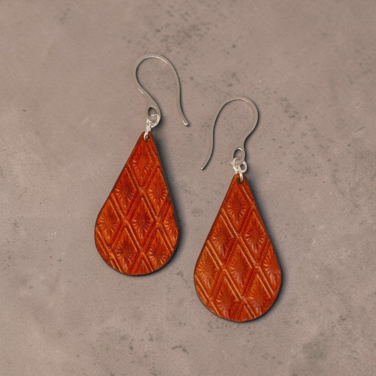Boucles d'oreilles forme goutte d'eau en cuir teinture cognac, estampages au matoir losange, vues de face, suspendues à des boucles argentées sur fond design