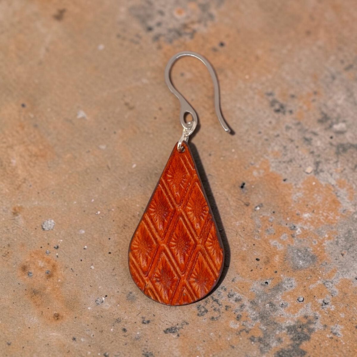 Boucle d'oreille forme goutte d'eau en cuir teinture cognac, estampage au matoir losange, vue de face, suspendue à une boucle argentée, fond design