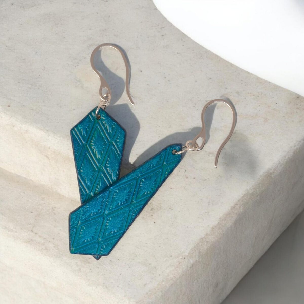 Boucles d'oreilles cravates asymétriques en cuir teinté bleu, estampage au matoir, vues de face se chevauchant, suspendues à des boucles argentées sur pierre