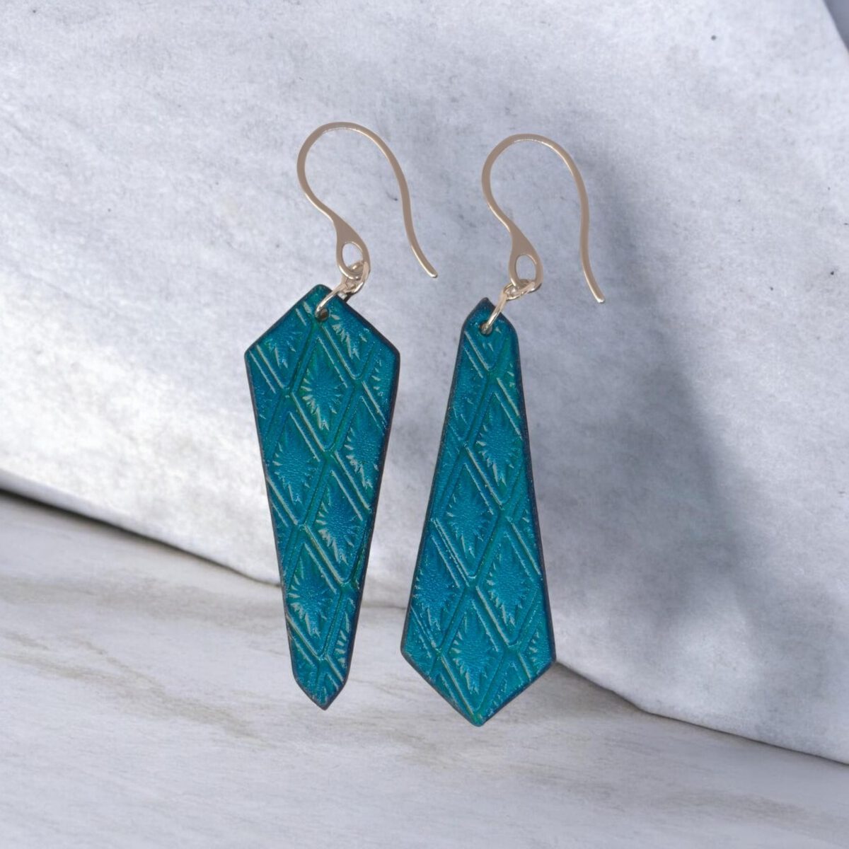 Boucles d'oreilles cravates asymétriques en cuir teinté bleu, estampage au matoir, vues de face, suspendues à des boucles argentées sur une pierre