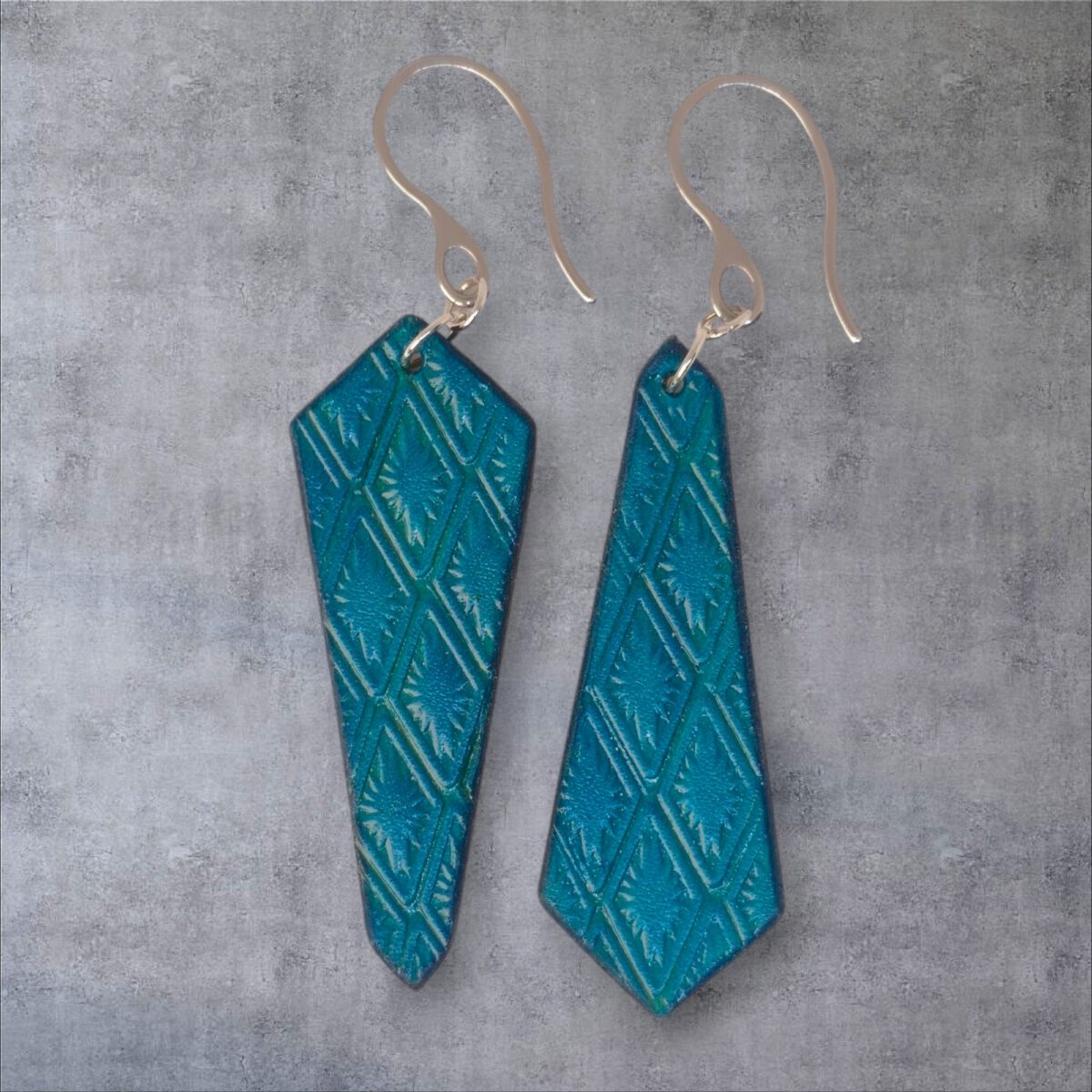 Boucles d'oreilles cravates asymétriques en cuir teinté bleu, estampage au matoir, vues de face, suspendues à des boucles argentées sur un fond gris