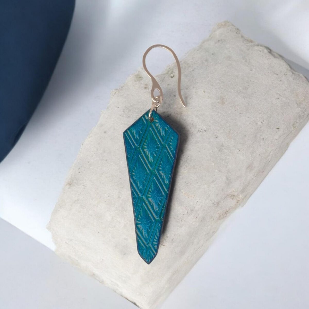 Boucle d'oreille cravate asymétrique en cuir teinté bleu, estampage au matoir, vue de face, suspendue à une boucle argentée sur pierre