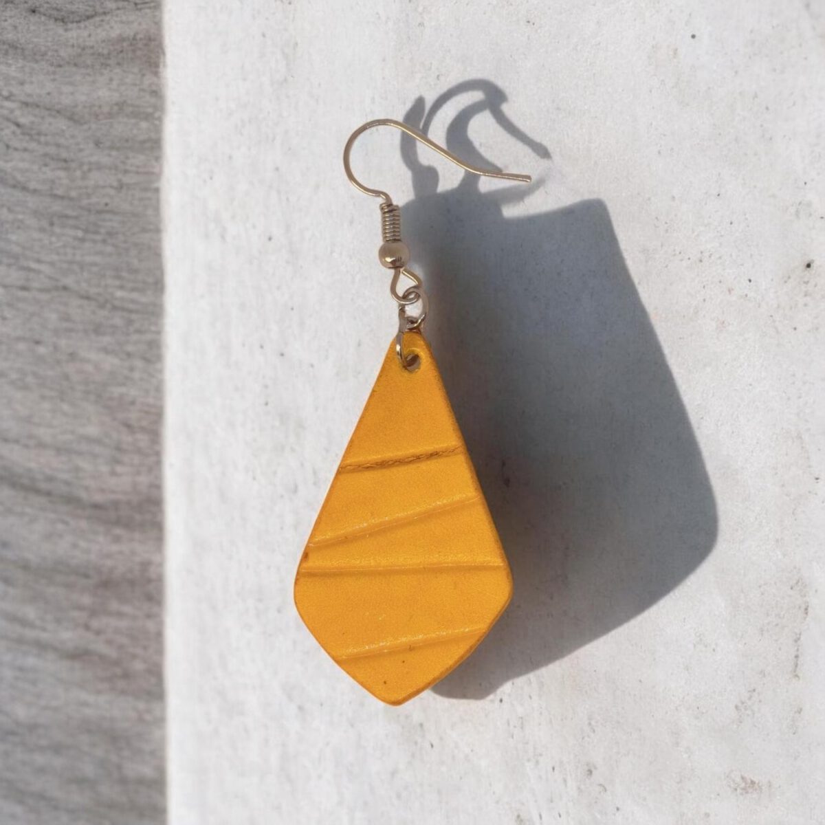 Boucle d'oreille forme larme en cuir teinture jaune, finition galet, vue de face, suspendue à une boucle argentée, sur pierre