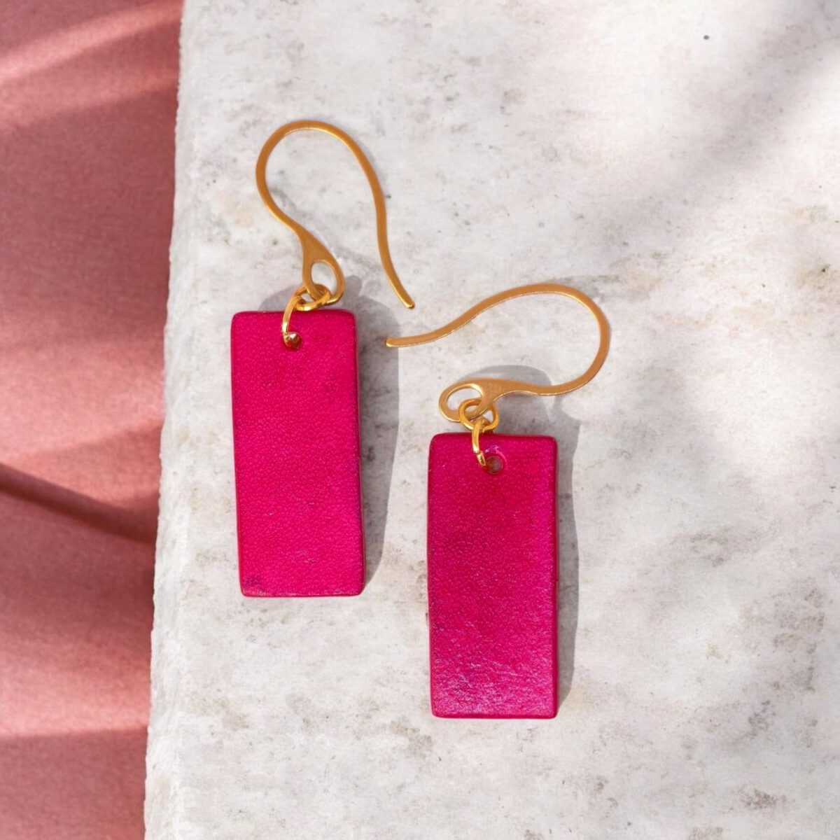 Boucles d'oreilles en cuir teintées en fuschia très girly et vives, vues de face, suspendues à des crochets dorés sur pierre