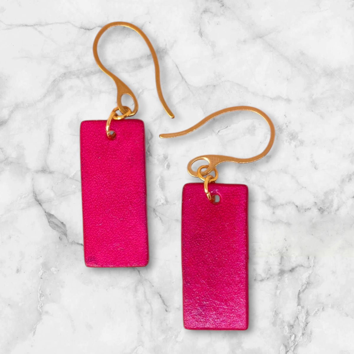 Boucles d'oreilles en cuir teintées en fuschia très girly et vives, vues de face, suspendues à des crochets dorés sur un fond marbré