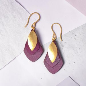 Boucles d'oreilles forme larme doublée en cuir teinture violette, finition lisse et pendant poisson doré brossé, vues de face, suspendues à des boucles dorées sur fond design